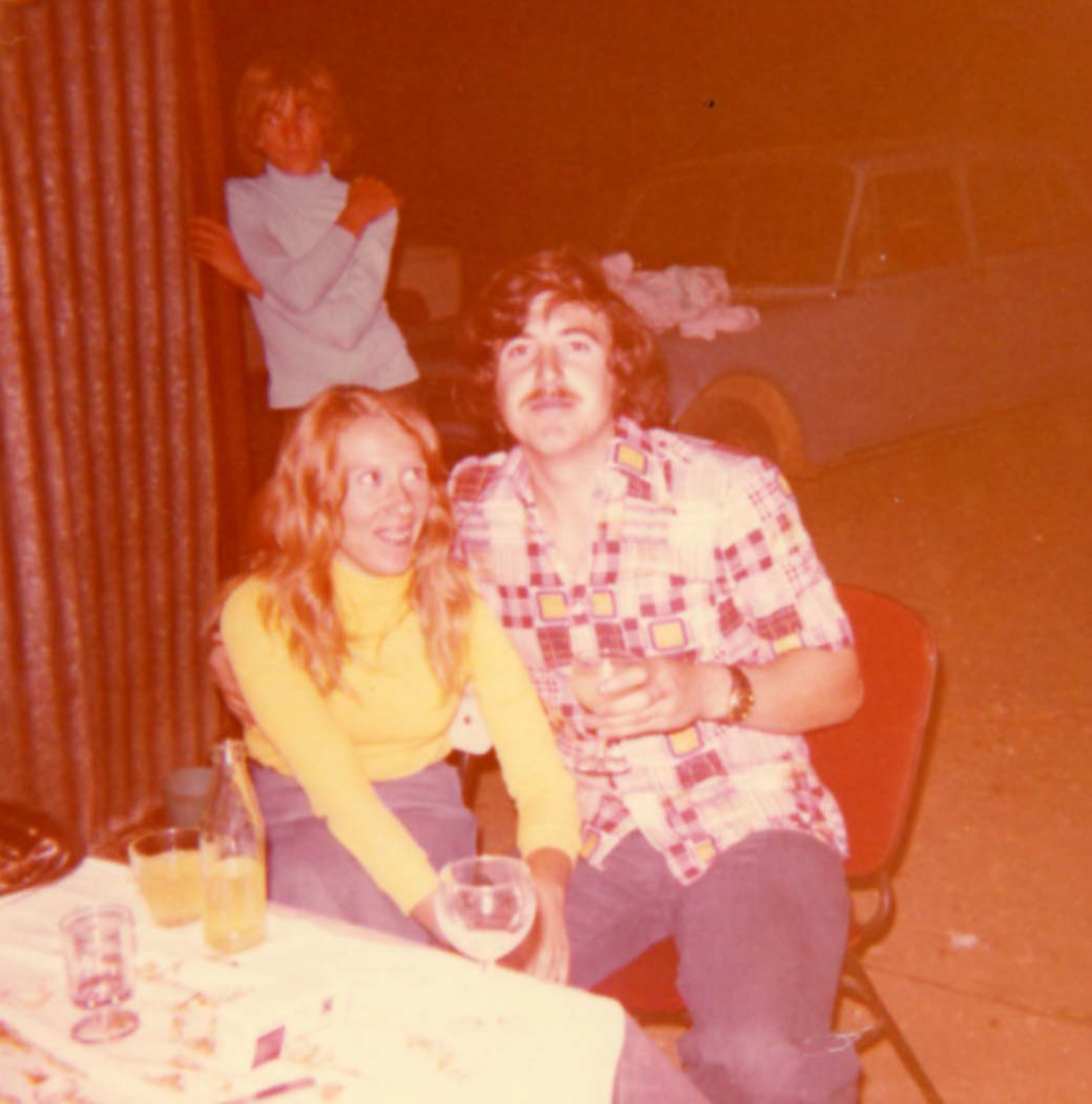 Une photographie d'époque montre un homme et une femme assis à une table, probablement dans les années 1970. L'homme a une moustache et porte une chemise à carreaux colorée, tandis que la femme est vêtue d'un pull jaune. Ils tiennent tous les deux des boissons. Une autre femme se tient à l'arrière-plan, portant un haut bleu clair, les mains près du visage. Cette charmante scène capturée par photographe de baptême