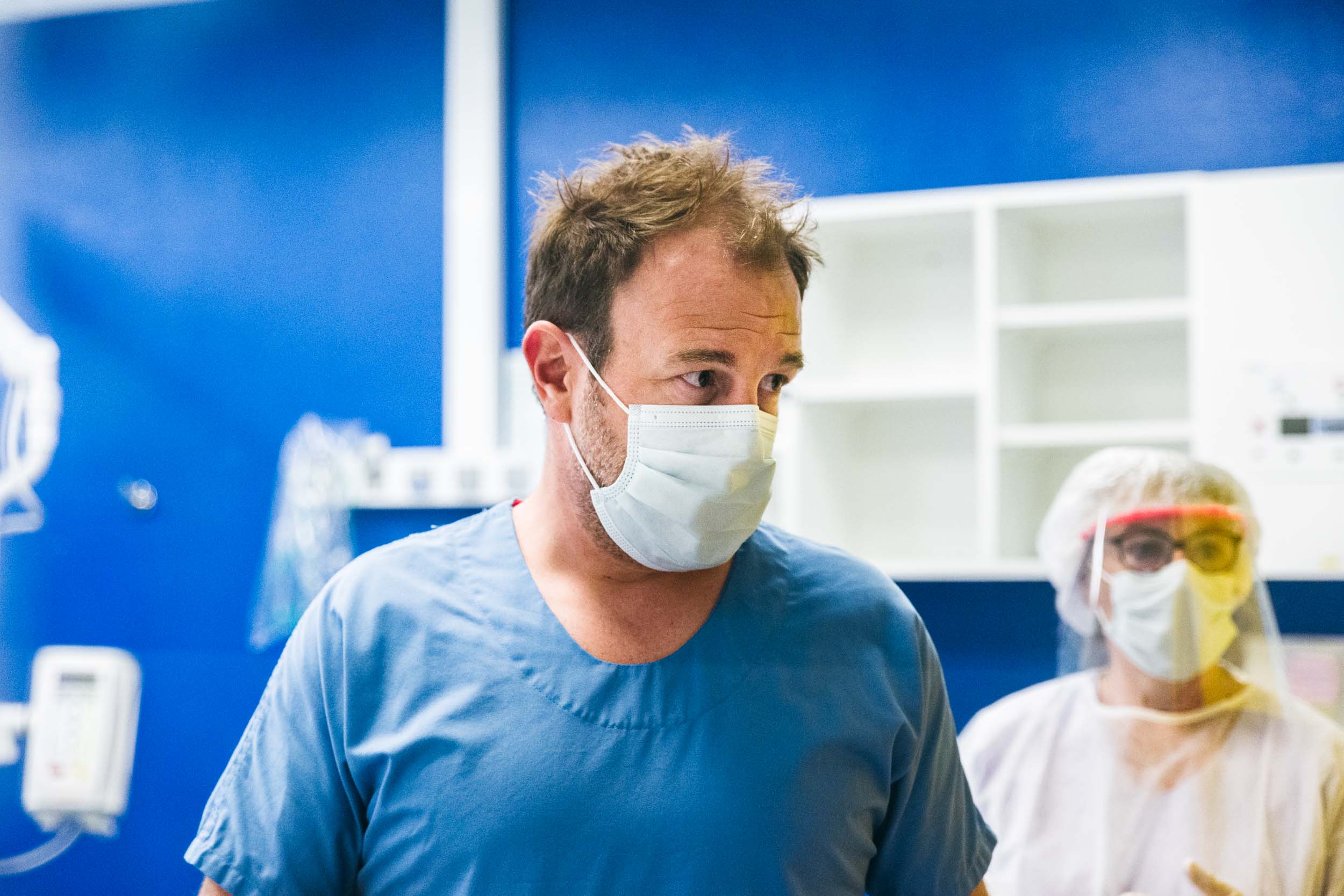 Un soignant, vêtu d'une tenue médicale bleue et d'un masque, se tient debout dans un cadre clinique aux murs bleu vif et aux étagères blanches. Derrière lui, un autre soignant, portant un équipement de protection complet, comprenant un masque facial, des lunettes et un couvre-cheveux, est partiellement visible. Il s'agit de véritables Héros Méconnus du CH d'Angoulême.