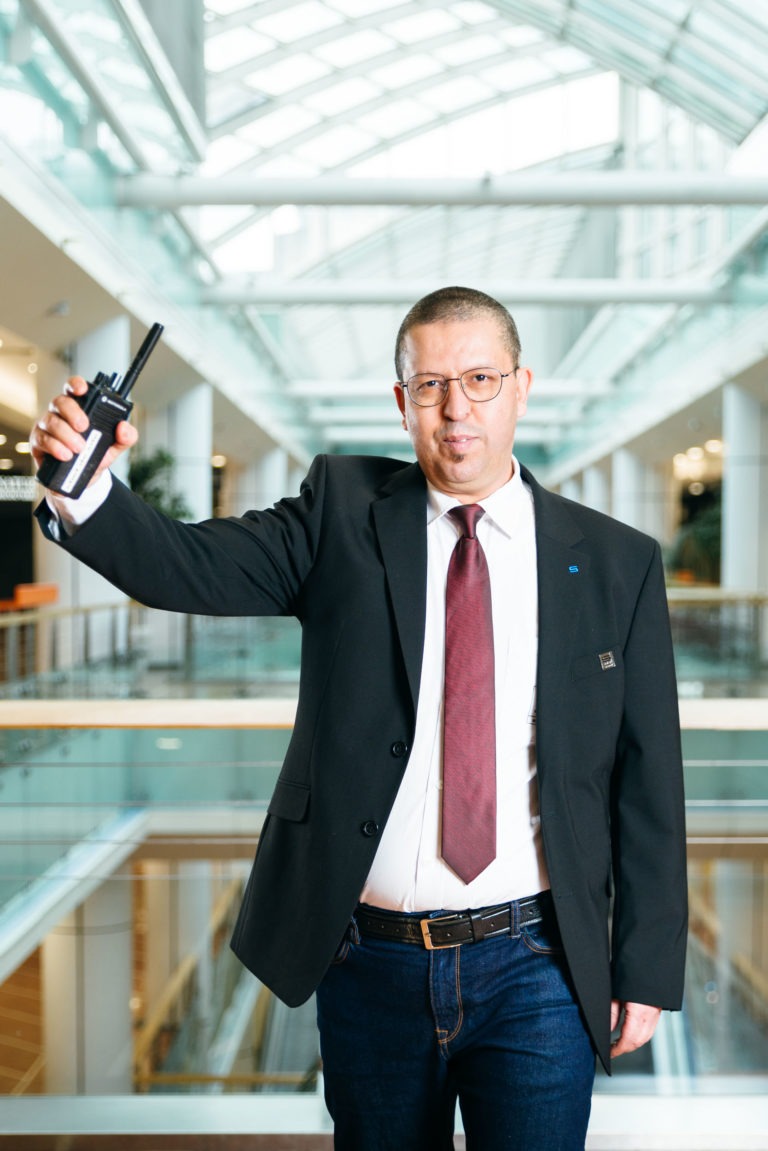 Un homme portant des lunettes, un blazer noir, une chemise blanche, une cravate rouge et un jean se tient debout à l'intérieur de l'atrium moderne de l'Hyper-Héros Centre Commercial Mériadeck. Il tient un talkie-walkie dans sa main droite et regarde directement la caméra. L'arrière-plan présente un bâtiment à plusieurs niveaux avec des balustrades et un éclairage.