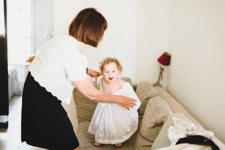 Un enfant vêtu d'une robe blanche en dentelle se tient debout sur un canapé beige, l'air joyeux et excité. Un adulte, vêtu d'un haut blanc et d'une jupe sombre, aide l'enfant, l'aidant peut-être à préparer la robe pour leur simple mariage champêtre. La pièce est lumineuse, la lumière pénètre par une fenêtre et une petite lampe rouge décore l'arrière-plan.