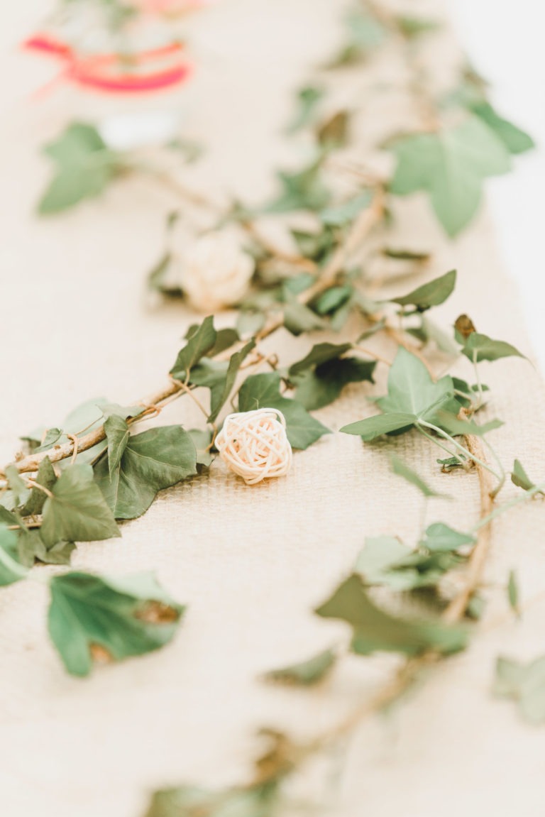 Gros plan d'une composition décorative composée d'une vigne aux feuilles vertes et d'une petite boule beige finement tissée, placée sur une surface en tissu texturé. L'arrière-plan est légèrement flou, capturant l'essence d'un mariage champêtre simple et mettant en valeur les détails de la vigne et de la boule tissée au premier plan.