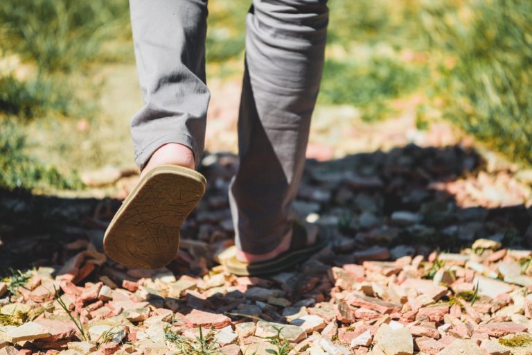 Une personne portant un pantalon gris et des tongs vertes marche sur un chemin rocailleux entouré d'herbe. L'image est centrée sur les jambes et les pieds de la personne, capturant le mouvement de la marche par derrière. La lumière du soleil projette des ombres naturelles sur le sol irrégulier et parsemé de rochers.