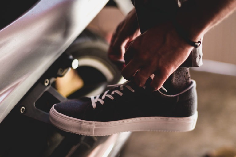 Gros plan d'une personne qui noue les lacets d'une basket noire à semelles blanches, tout en posant son pied sur un repose-pied de moto. L'individu porte un pantalon sombre et un bracelet au poignet. La scène semble aussi intime et concentrée que les préparatifs d'un simple mariage champêtre.