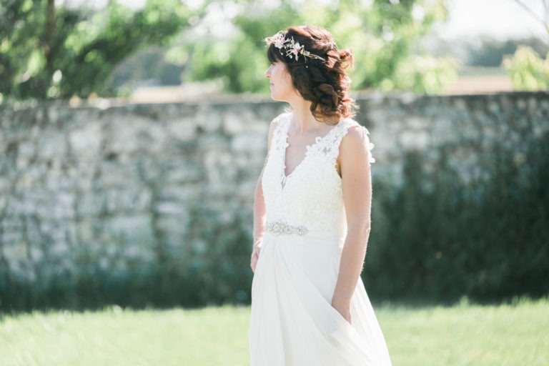 Une mariée se tient debout devant un mur de pierre, à l'extérieur, dégageant le charme d'un mariage champêtre simple. Elle porte une robe de mariée en dentelle blanche avec un corsage en dentelle florale et une ceinture en satin. Ses cheveux sont coiffés en boucles lâches ornées d'un délicat couvre-chef floral, tandis que l'arrière-plan présente une verdure floue et la lumière du soleil.