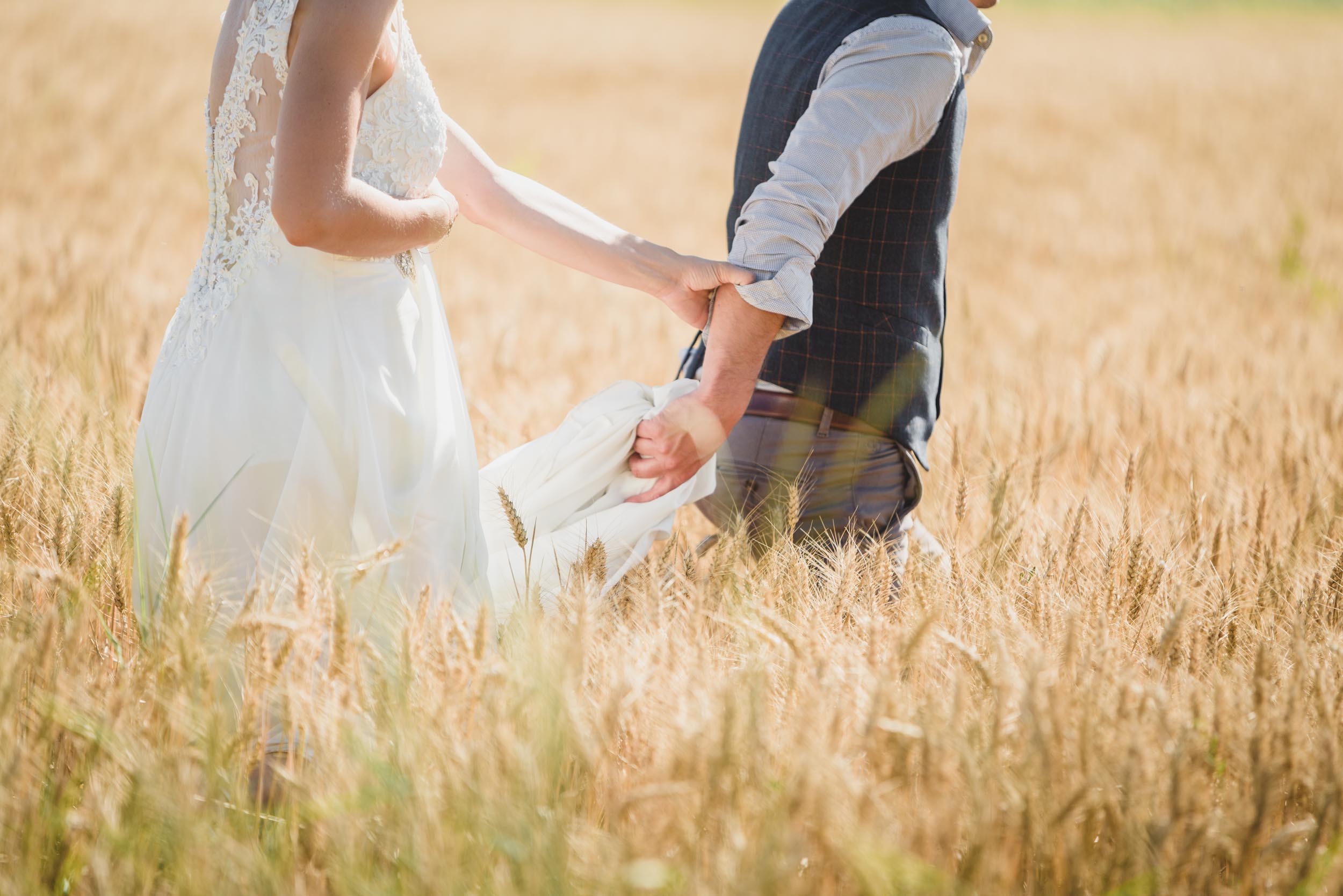 Un couple de mariés marche main dans la main dans un champ de blé doré. La mariée porte une robe de mariée en dentelle blanche et le marié est habillé d'un gilet et de manches retroussées. Ils sont photographiés en plein pas, entourés de tiges de blé dorées sous un ciel clair et ensoleillé, incarnant l'essence d'un mariage champêtre simple.