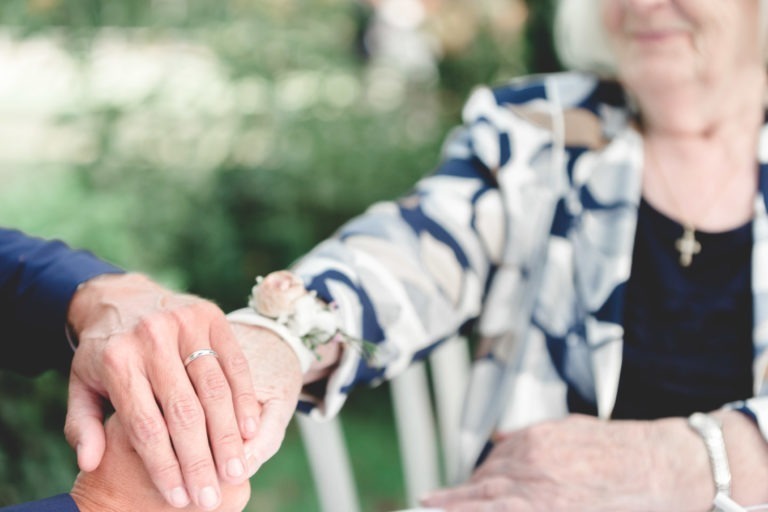 Une femme âgée, vêtue d'un blazer bleu marine et blanc à fleurs, pose doucement sa main sur celle d'une autre femme, qui porte une alliance. L'arrière-plan est luxuriant et vert, suggérant un décor extérieur. Le gros plan souligne la simplicité et l'affection de ce geste tendre de connexion.