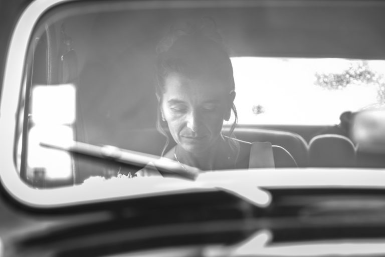 Une photographie en noir et blanc montre une femme assise dans une voiture, vue à travers le pare-brise. Sa tête est penchée et elle semble contemplative, une douce lumière éclairant son visage. L'essuie-glace flou est visible au premier plan, ajoutant une texture subtile à l'image, évoquant la simplicité et l'introspection.