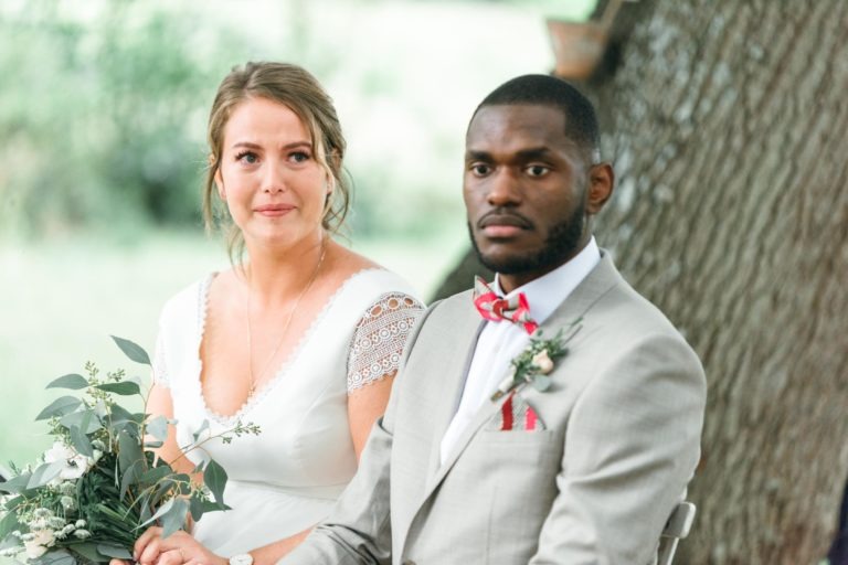 Une mariée et un marié sont assis côte à côte à l'extérieur. La mariée, dans une robe blanche aux manches en dentelle, tient un bouquet vert et blanc et porte un collier pendentif. Le marié, dans un costume gris clair avec un nœud papillon rouge et une pochette, a une boutonnière sur son revers. Tous deux regardent devant eux avec des expressions calmes, leur mariage émouvant et touchant.