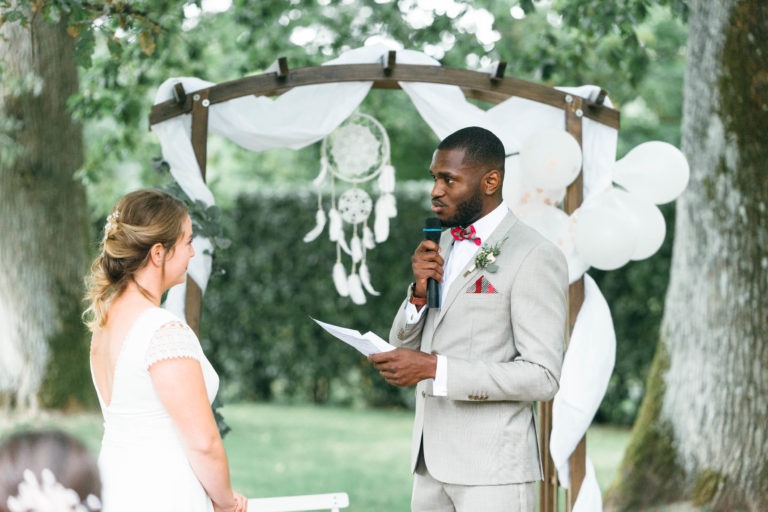 Un couple de mariés se tient face à face sous une arche en bois décorée de tissu blanc et d'attrape-rêves. Le marié, en costume gris clair avec un nœud papillon rouge, tient un micro et un papier, lisant vraisemblablement des vœux. La mariée porte une robe blanche. Des arbres et de la verdure entourent ce mariage émouvant et touchant.