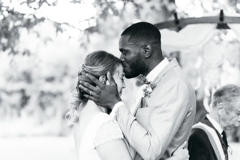 Une photo en noir et blanc capture un moment émouvant et touchant lors d'un mariage. Un marié embrasse doucement le front de la mariée tout en tenant son visage avec ses deux mains. Le couple se tient sous une arche ornée de décorations florales, et un officiant est visible en arrière-plan.