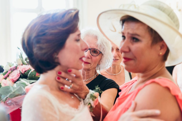 Trois femmes partagent un moment d'émotion lors d'un mariage. La mariée, en robe blanche, est délicatement tenue par une femme en robe rose. Toutes deux semblent avoir les yeux pleins de larmes. Une femme âgée aux cheveux blancs et aux lunettes à l'arrière-plan regarde, visiblement émue par cette renaissance émotionnelle. Des fleurs roses ornent l'arrière-plan à gauche.