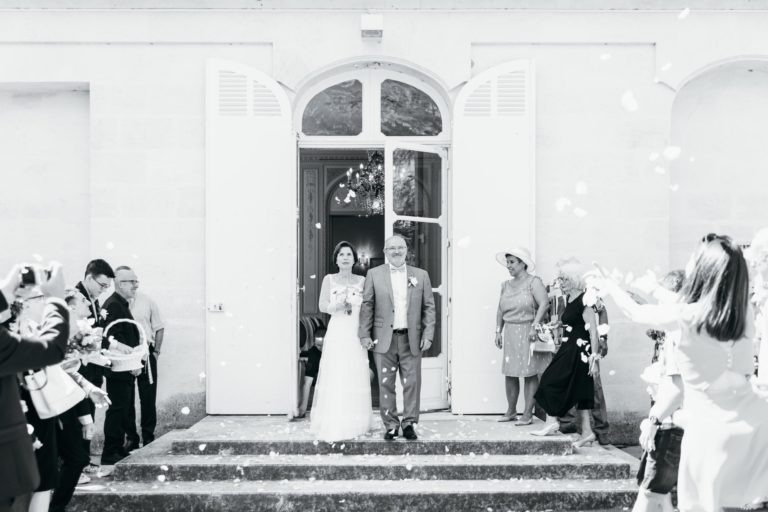 Une mariée et un homme plus âgé se tiennent côte à côte en haut des marches devant des portes doubles. Ils sont entourés de personnes qui lancent des pétales de fleurs. La mariée, tenant un bouquet et souriant, dégage une émotion de renaissance, capturant le moment joyeux à la fin d'une cérémonie de mariage.