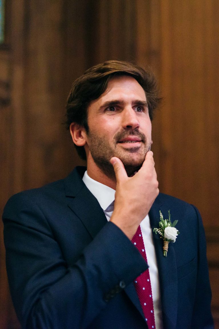 Un homme aux cheveux bruns et à la barbe est vêtu d'un costume sombre avec une chemise blanche et une cravate à pois rouges. Touchant son menton avec sa main droite, il regarde légèrement vers le haut. Une petite boutonnière florale est épinglée à sa veste de costume. Le fond en boiseries ajoute une touche élégante, parfaite pour un mariage Parfait à Bordeaux.