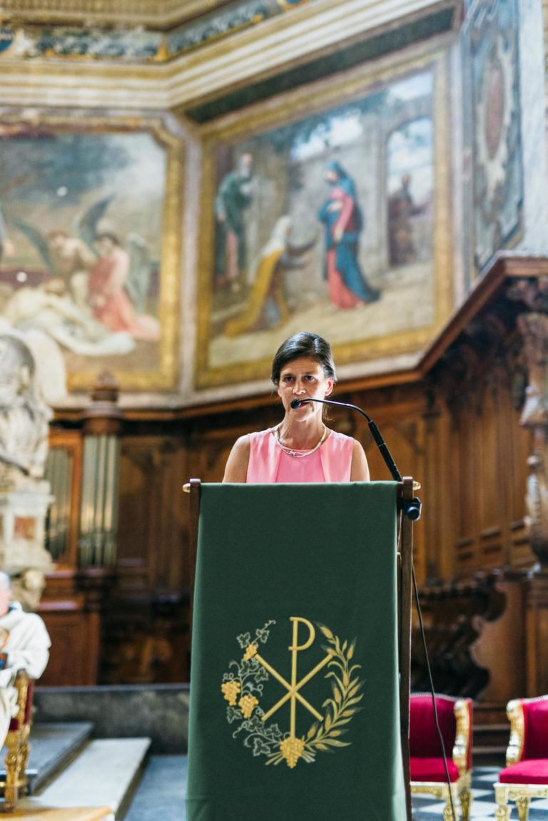 Une femme vêtue d'un haut rose sans manches parle dans un micro sur un podium drapé d'un tissu vert sur lequel est apposé un symbole religieux doré. L'arrière-plan présente des panneaux de bois ornés et des œuvres d'art religieuses détaillées sur les murs. Des chaises rouges sont visibles sur le côté, plantant le décor du mariage Parfait à Bordeaux.