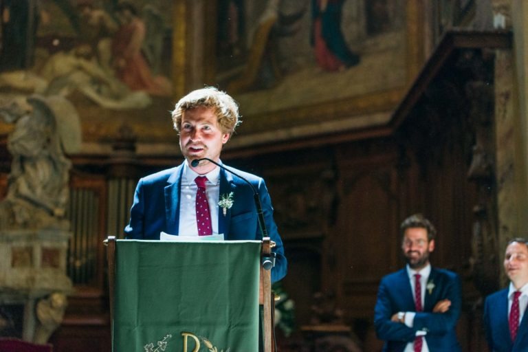 Un homme en costume se tient debout sur une estrade avec un micro, prononçant un discours à l'intérieur. Derrière lui, deux hommes en costume écoutent attentivement, dont l'un sourit. La salle est décorée de sculptures et de peintures rappelant un Mariage Parfait à Bordeaux. La tribune est partiellement recouverte d'un tissu vert.