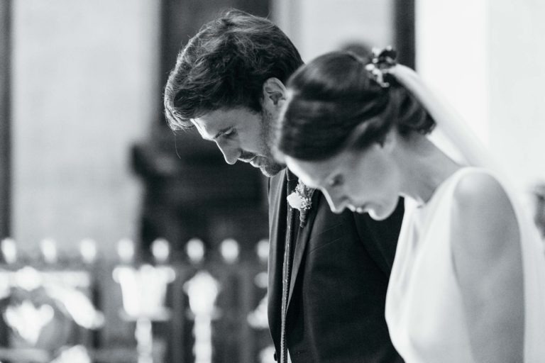 Une photo en noir et blanc montre un couple de mariés debout côte à côte dans un moment de réflexion lors de leur mariage Parfait à Bordeaux. Le marié, vêtu d'un costume, et la mariée, vêtue d'une robe blanche et d'un voile, ont tous deux la tête penchée. L'arrière-plan est légèrement flouté.