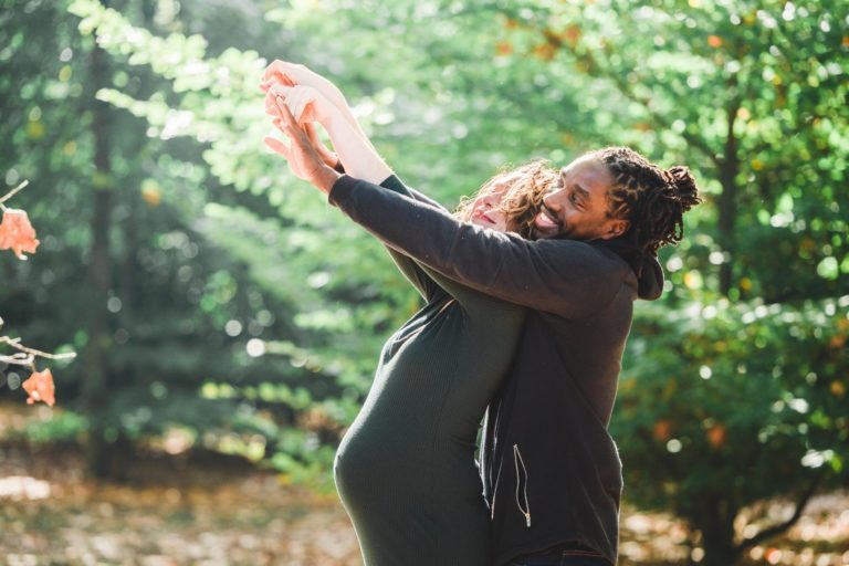 Une femme enceinte en robe verte et un homme en sweat à capuche noir se tiennent ensemble à l'extérieur. Il se tient derrière elle, la serrant fort dans ses bras avec un sourire joyeux. Elle lève les bras de manière enjouée vers les feuilles orange, tandis que la lumière du soleil filtre à travers les arbres verts et luxuriants, capturant la beauté de leur séance photo intime entre mère et fille.