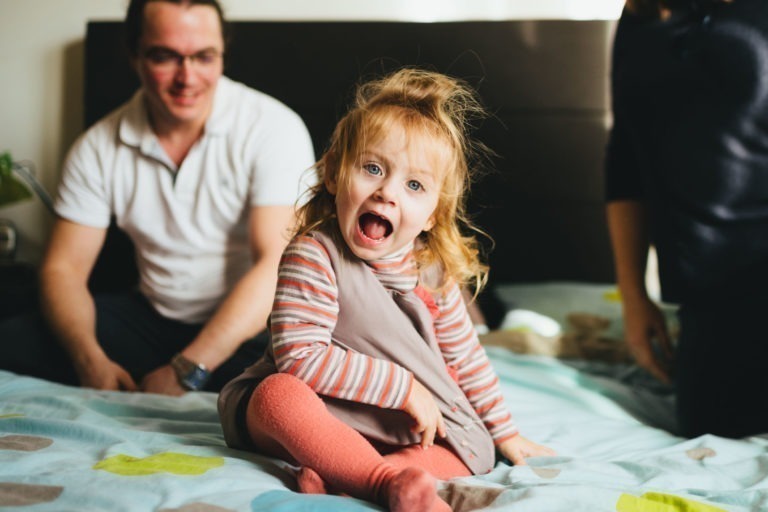 Un jeune enfant joyeux aux cheveux châtain clair est assis sur un lit, vêtu d'un tablier gris sur une chemise rayée et d'un legging corail. La bouche de l'enfant est grande ouverte d'excitation. Un adulte souriant en chemise blanche, appréciant probablement l'atmosphère chaleureuse de la Maison du Bonheur, est assis derrière l'enfant, et une autre personne est partiellement visible sur la droite.