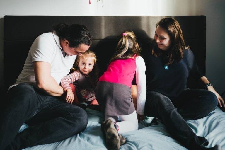 Une famille de quatre personnes est assise sur un lit au milieu de la lumière naturelle. La mère et le père, habillés de façon décontractée, sont assis de chaque côté à la Maison du Bonheur. Le père chatouille une petite fille blonde en chemise rayée qui rigole. Un enfant plus âgé, le dos tourné, est penché en avant, vêtu d'un haut rose et d'une jupe grise. La mère sourit chaleureusement.