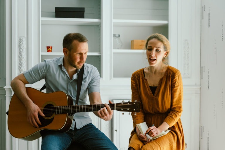 Un homme et une femme, connus sous le nom du duo Altalina, font de la musique dans une pièce lumineuse aux murs et aux étagères blancs. L'homme, vêtu d'une chemise bleu clair et d'un jean, joue de la guitare acoustique. La femme, vêtue d'une robe couleur moutarde, tient une tasse blanche et chante. Ils semblent concentrés et engagés dans leur performance.