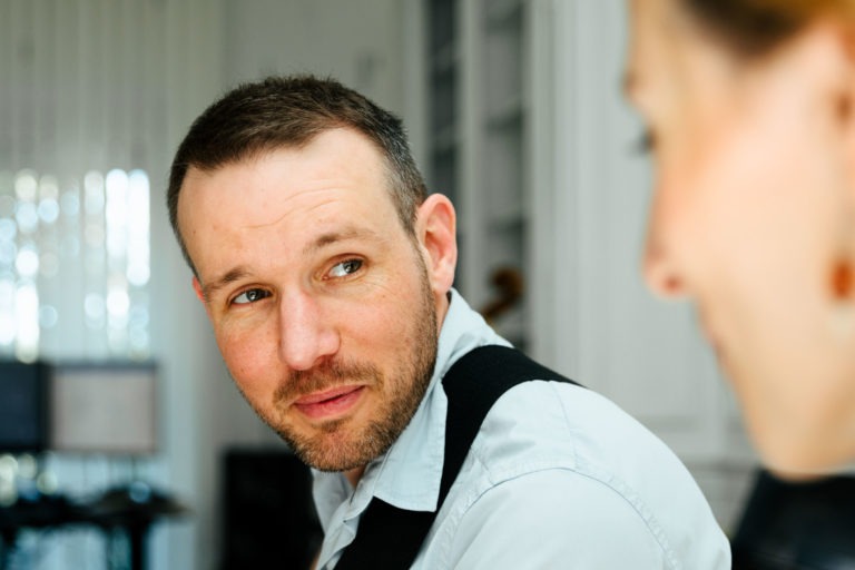 Un homme aux cheveux courts et à la barbe taillée, vêtu d'une chemise bleu clair et de bretelles noires, regarde attentivement une femme au premier plan dont le visage est flou. Ils semblent être dans un bureau bien éclairé avec des écrans d'ordinateur en arrière-plan, discutant peut-être d'un prochain événement musical du duo Altalina en Gironde.