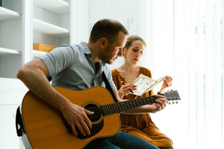 Un homme du duo Altalina joue de la guitare acoustique assis. A côté de lui, une femme en robe marron regarde un livre à la couverture fleurie, semblant suivre la musique. Ils sont à l'intérieur de leur maison girondine, avec des étagères blanches et une fenêtre avec des stores verticaux doucement éclairée par la lumière naturelle.