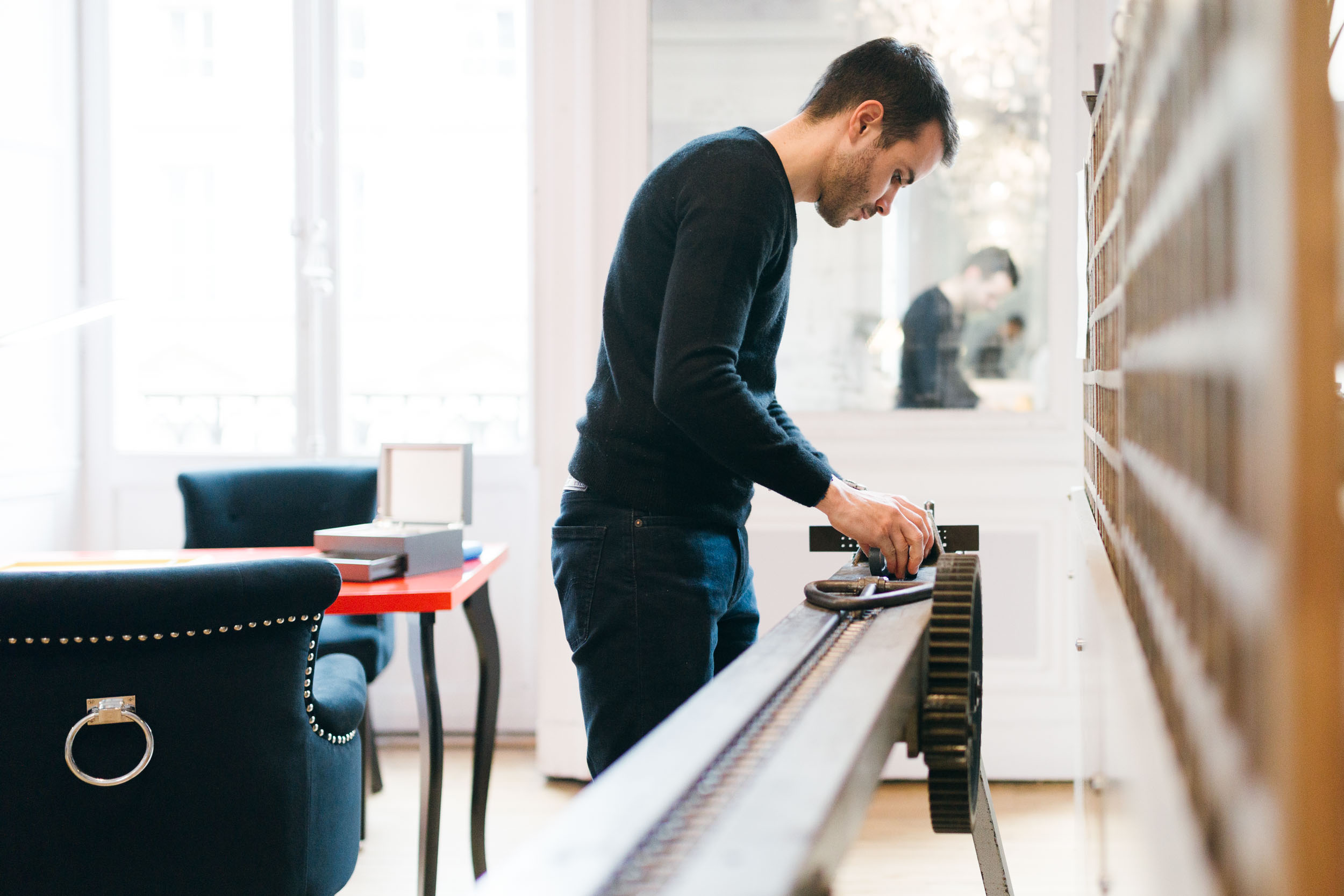 Dans l'Atelier du Serti, un homme vêtu d'un pull noir et d'un jean foncé travaille sur un long appareil mécanique. Il semble concentré sur sa tâche. La pièce est dotée de murs clairs, d'une chaise avec un dossier clouté et une poignée en anneau, et d'une table rouge à proximité. La grande fenêtre à l'arrière-plan laisse entrer la lumière naturelle.