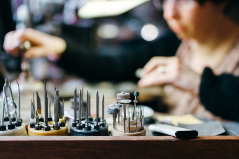 Un gros plan de l'atelier de bijouterie de l'Atelier du Serti présente divers outils spécialisés disposés dans des supports sur un banc en bois. La silhouette floue d'une personne à l'arrière-plan s'attaque à une pièce de métal ou à un bijou, suggérant un travail minutieux. La scène met l'accent sur la précision et l'attention aux détails.