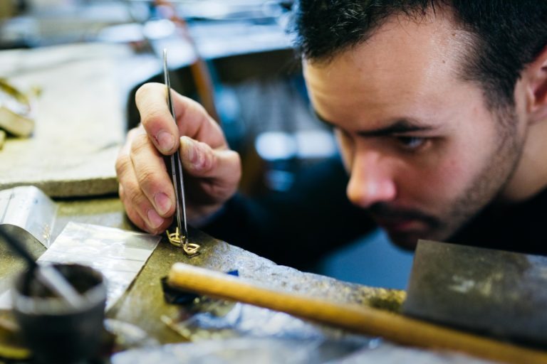 Gros plan sur une personne qui se concentre sur un travail artisanal complexe à l'Atelier du Serti. Elle utilise une pince pour manipuler de petits composants dorés sur un établi. Divers outils et matériaux, dont un petit marteau et une loupe, sont éparpillés un peu partout. L'arrière-plan est flou, ce qui met en valeur la concentration.