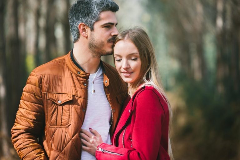 Un couple se tient dans une étreinte affectueuse dans une forêt, dans le cadre de leur séance photo en amoureux à Bordeaux. L'homme, vêtu d'une veste marron, penche la tête vers la femme, vêtue d'une veste rouge, les yeux fermés et un sourire satisfait. L'arrière-plan est flou, rempli de grands arbres, suggérant un cadre paisible et naturel.