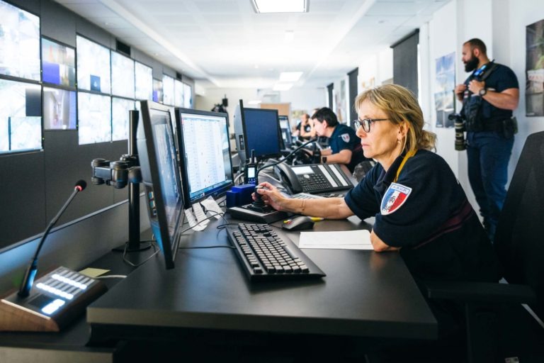 Une femme aux cheveux courts et aux lunettes travaille à un bureau avec plusieurs écrans d'ordinateur dans une salle de contrôle de surveillance. Elle porte un uniforme bleu marine avec un écusson brodé de la Police Municipale de Bordeaux. D'autres membres du personnel, dont un avec un équipement vidéo, sont également présents dans la pièce remplie de grands écrans.