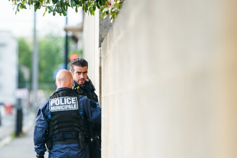 Deux policiers en uniforme bleu se tiennent debout près d'un mur. L'un d'eux, tourné vers l'extérieur, porte l'inscription « Police Municipale » au dos de son gilet. L'autre, tourné vers la caméra, semble engagé dans une conversation. Un feuillage vert est suspendu au-dessus d'eux sur le côté gauche de l'image. Leurs uniformes portent l'inscription « Police Municipale de Bordeaux ».