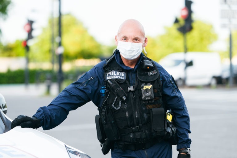 Un policier municipal de Bordeaux portant un masque et vêtu d'un uniforme bleu marine avec divers équipements se tient à l'extérieur près d'un véhicule. Il a le crâne chauve et tient la voiture avec sa main droite. L'arrière-plan présente des feuilles vertes, des feux de circulation et une camionnette blanche.