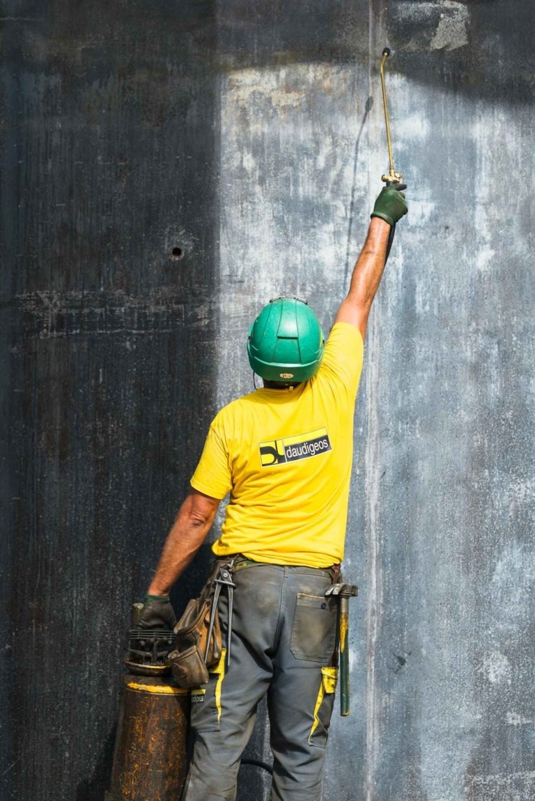 Un ouvrier du bâtiment portant un casque de chantier vert et une chemise jaune avec le logo « Daudigeos entreprise de gros-œuvre » peint une grande surface verticale avec une peinture sombre. Debout sur le côté gauche, son bras droit est tendu vers le haut, tenant la buse de pulvérisation. Il porte l'équipement sur une ceinture autour de sa taille.