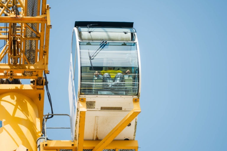 Une nacelle en verre d'une grande roue, avec une personne assise à l'intérieur en chemise jaune, est photographiée sur fond de ciel bleu clair. La structure jaune de la grande roue de l'entreprise de gros-œuvre Daudigeos est visible sur le côté gauche de l'image.