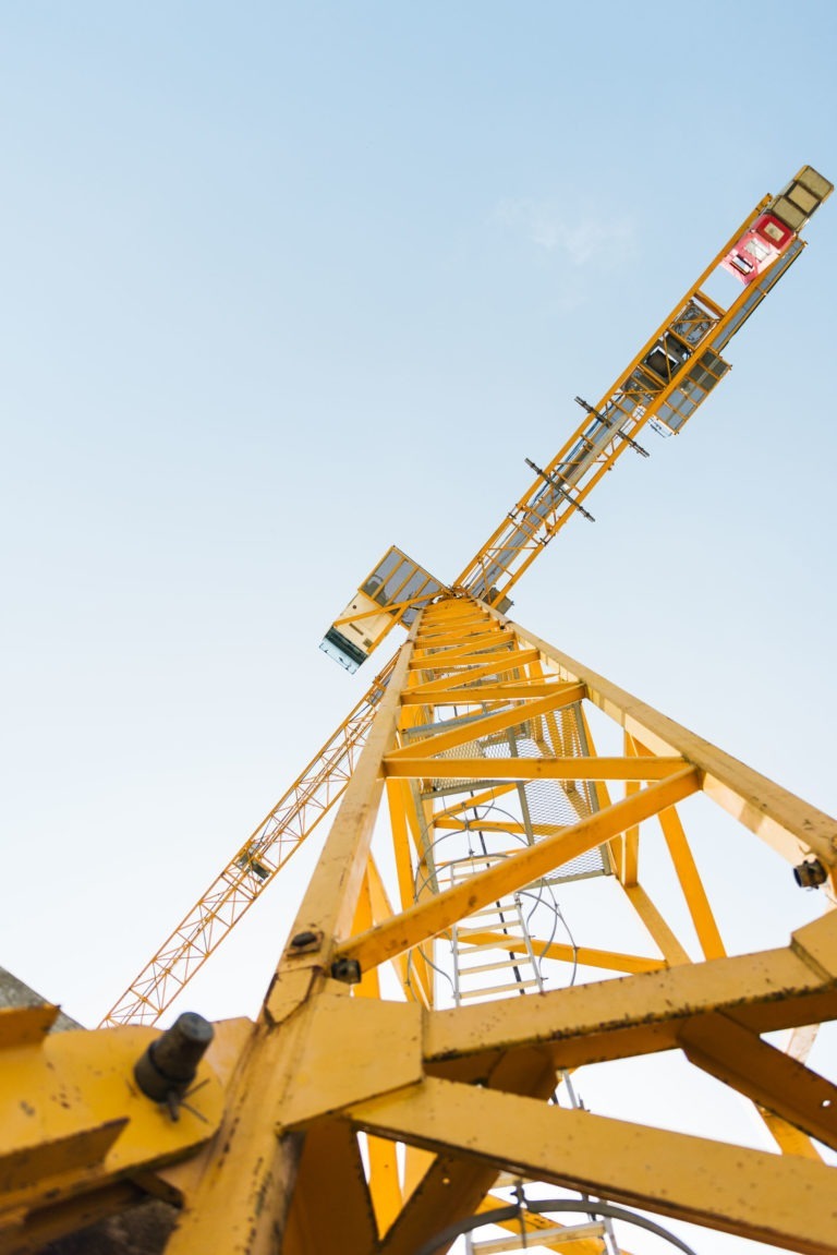 Vue en contre-plongée d'une grande grue de chantier jaune appartenant à l'entreprise de gros-œuvre Daudigeos sur fond de ciel bleu clair. La structure de la grue est bien mise en valeur, montrant sa structure complexe et son échelle. L'image capture l'ampleur et la hauteur de la grue alors qu'elle monte dans le ciel.