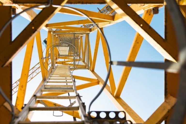 Vue directe vers le haut à l'intérieur d'une structure de grue en métal jaune avec des poutres interconnectées par l'entreprise de gros-œuvre Daudigeos. Le ciel bleu est visible à travers les espaces entre les poutres. Il y a une échelle qui monte verticalement au centre, avec des arceaux de sécurité circulaires qui l'entourent pour la protection.