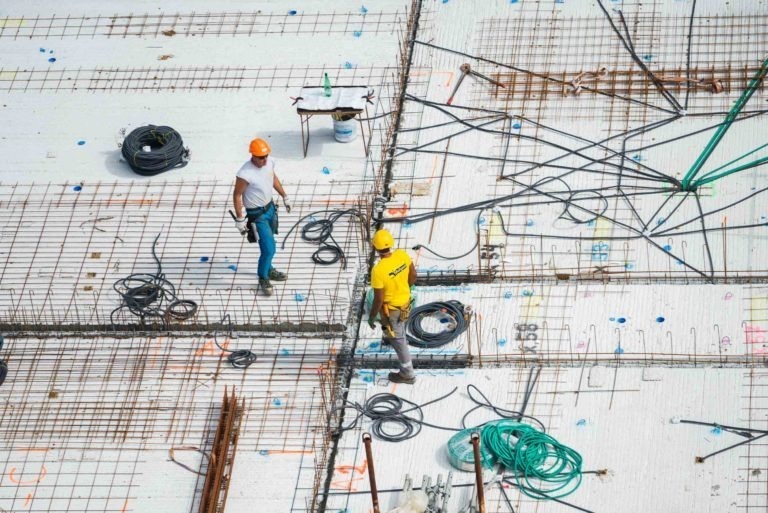 Deux ouvriers du bâtiment de l'entreprise de gros-œuvre Daudigeos, l'un en gilet jaune et l'autre en chemise blanche, travaillent sur un chantier de construction avec une charpente en acier. Des câbles enroulés, des outils et des matériaux de construction sont éparpillés un peu partout. Les ouvriers portent des casques et des équipements de sécurité.