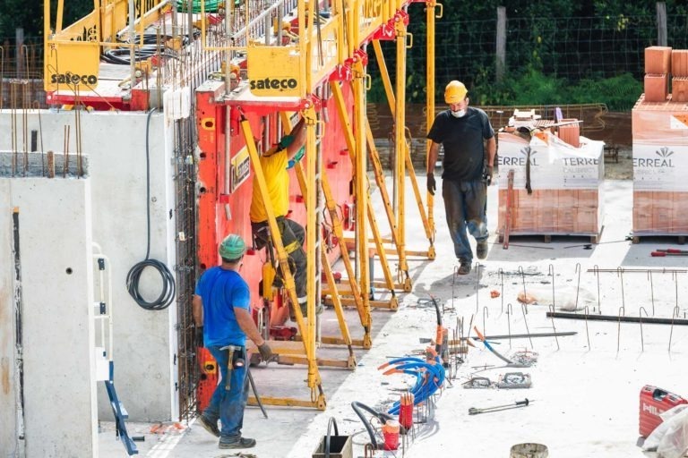 Trois ouvriers du bâtiment travaillent sur un chantier en activité de l'entreprise de gros-œuvre Daudigeos. Un ouvrier en jaune monte sur un échafaudage, un autre en bleu est au sol avec des outils et le troisième en noir marche sur une surface en béton. Divers matériaux et équipements de construction sont éparpillés sur le chantier.