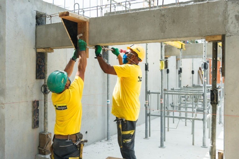 Deux ouvriers du bâtiment portant des chemises jaunes, des casques de chantier et des gants travaillent sur une structure en béton sur un chantier de construction pour l'entreprise de gros-œuvre Daudigeos. L'ouvrier de gauche tient une fixation en bois tandis que l'ouvrier de droite la fixe à l'aide d'un marteau. Le chantier est entouré d'échafaudages et de piliers en béton.