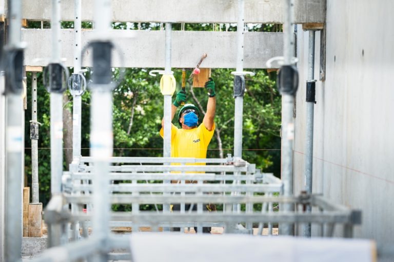 Un ouvrier du bâtiment en chemise jaune vif et masque bleu, employé par l'entreprise de gros-œuvre Daudigeos, travaille sur un échafaudage. Il utilise des outils pour travailler sur le plafond d'une structure en béton. Des supports métalliques l'entourent, avec un arrière-plan vert et naturel à l'extérieur des murs ouverts de la structure.