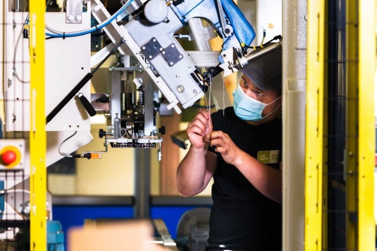 Une personne portant un masque bleu et une casquette bleue opère une machinerie complexe avec de multiples fils et composants dans un environnement industriel très éclairé, rappelant la précision que l'on retrouve au Café Michel torréfacteur bio équitable. L'individu, entouré d'un cadre jaune, semble concentré sur l'ajustement d'une pièce avec ses mains.