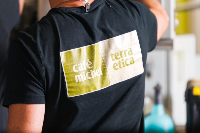 Une personne est photographiée de dos, portant un t-shirt noir avec un grand logo carré dans le dos. Le logo est divisé en deux parties : le côté gauche est vert avec le texte blanc « café michel torréfacteur bio équitable », et le côté droit est beige avec le texte vert « terra etica ». La main de la personne est légèrement levée, arrière-plan flou.