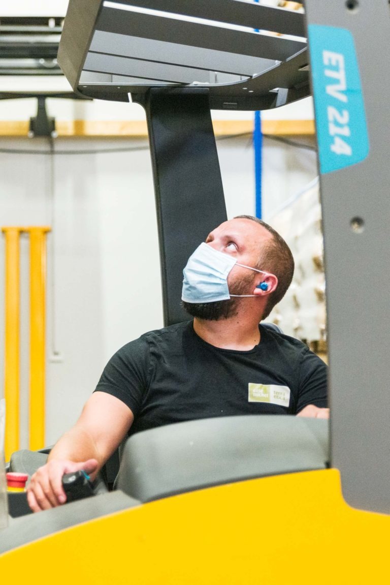 Un homme barbu, portant un masque et des bouchons d'oreilles bleus, conduit un chariot élévateur jaune étiqueté « ETV 214 » dans une installation intérieure. Le regard vers le haut, peut-être vers la charge, il passe devant des étagères et des équipements de sécurité. La scène rappelle le soin méticuleux observé au Café Michel torréfacteur bio équitable.