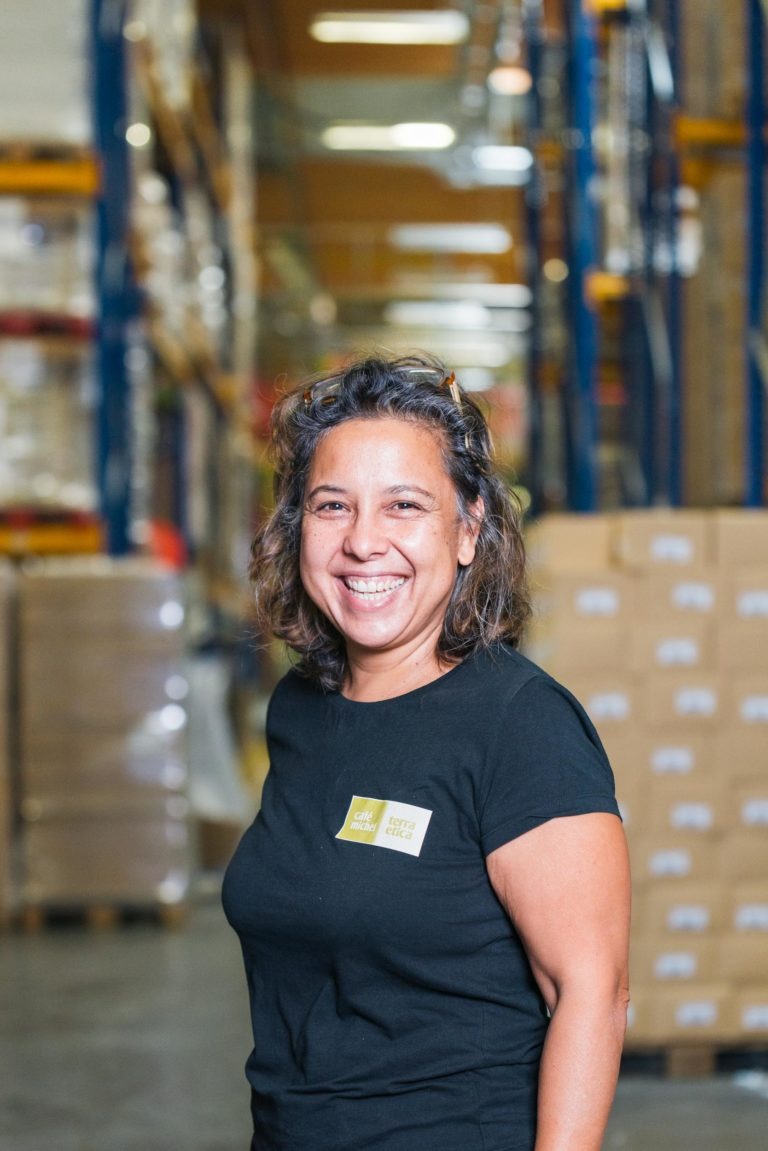 Une personne souriante, vêtue d’un t-shirt noir, se tient dans une allée d’entrepôt bordée de hautes étagères remplies de boîtes. L’arrière-plan est flou, mettant en valeur des rangées d’étagères remplies de colis, suggérant un espace de stockage animé et organisé. Peut-être vient-elle de recevoir des grains de café frais du Café Michel torréfacteur bio équitable.