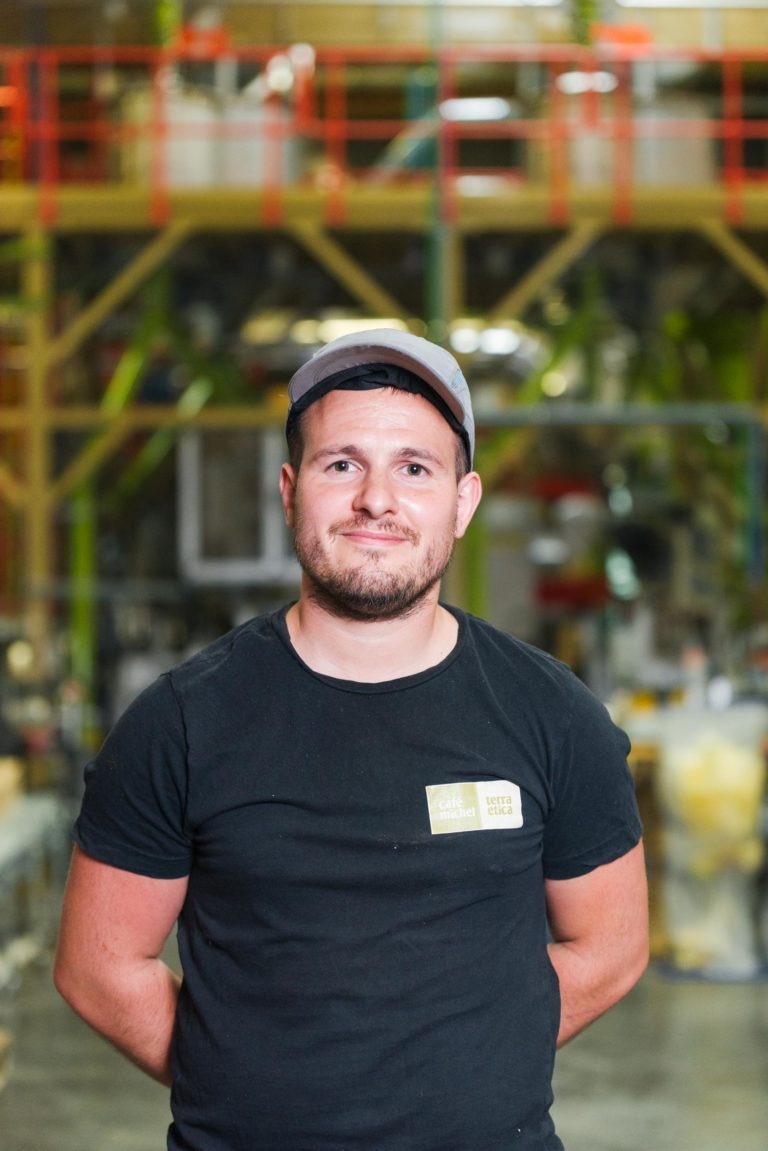 Un homme aux cheveux courts et à la barbe taillée, vêtu d’un t-shirt noir et d’une casquette claire, se tient debout, les mains derrière le dos. Il se trouve à l’intérieur, dans un décor d’atelier ou d’usine, rappelant les espaces animés où prennent vie les produits de qualité du Café Michel torréfacteur bio équitable. Des étagères et des équipements industriels sont visibles en arrière-plan flou.