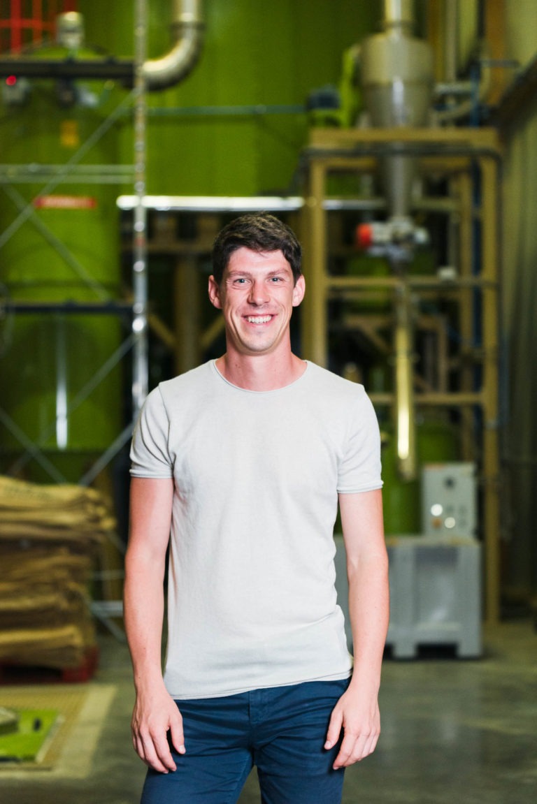 Une personne souriante, vêtue d'un t-shirt gris clair et d'un pantalon foncé, se tient debout dans un décor industriel. L'arrière-plan, qui rappelle une installation de torréfacteur bio équitable du Café Michel, présente des machines vertes, des tuyaux en métal et des palettes en bois, créant un environnement suggérant une usine ou un entrepôt. Les mains de la personne reposent à ses côtés.