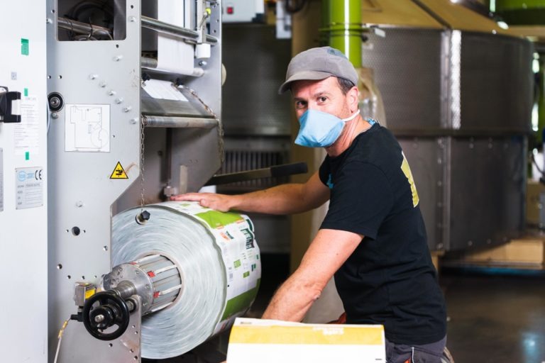 Une personne portant une casquette grise, une chemise noire et un masque bleu utilise une grande machine industrielle au Café Michel torréfacteur bio équitable. Elle ajuste un rouleau de matériau semblable à du plastique avec des étiquettes dessus. L'arrière-plan montre des machines et des équipements industriels, indiquant un environnement de fabrication animé.