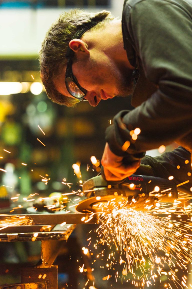 Une personne portant des lunettes de sécurité meule du métal avec une meuleuse d'angle dans l'atelier de la Ferronnerie d'Art de Bordeaux, produisant une pluie d'étincelles orange vif. Ils sont concentrés sur leur travail, avec des arrière-plans flous d'outils et d'équipements. La scène est éclairée par l'éclairage de l'atelier.