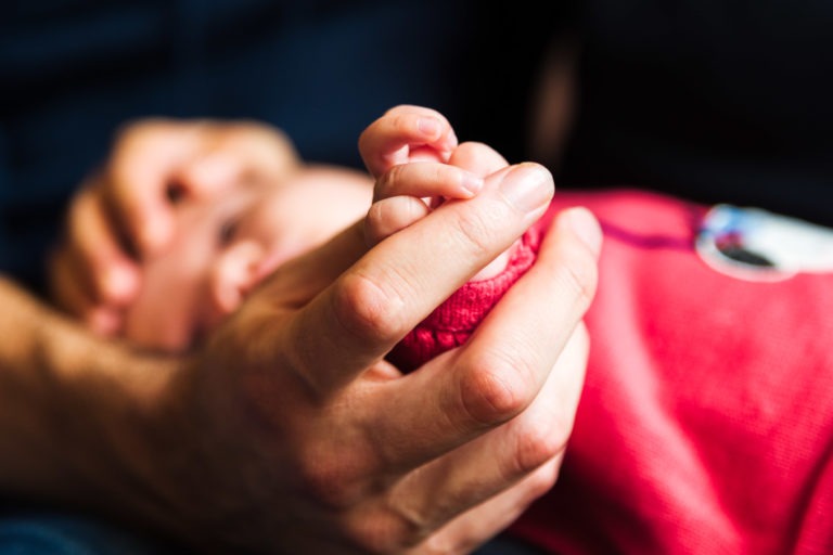 Gros plan de la main d'un adulte tenant doucement celle d'un bébé. Le bébé porte une tenue rouge et la scène capture un moment précieux de connexion et de soin en famille. L'arrière-plan est flou pour garder l'accent sur les mains.